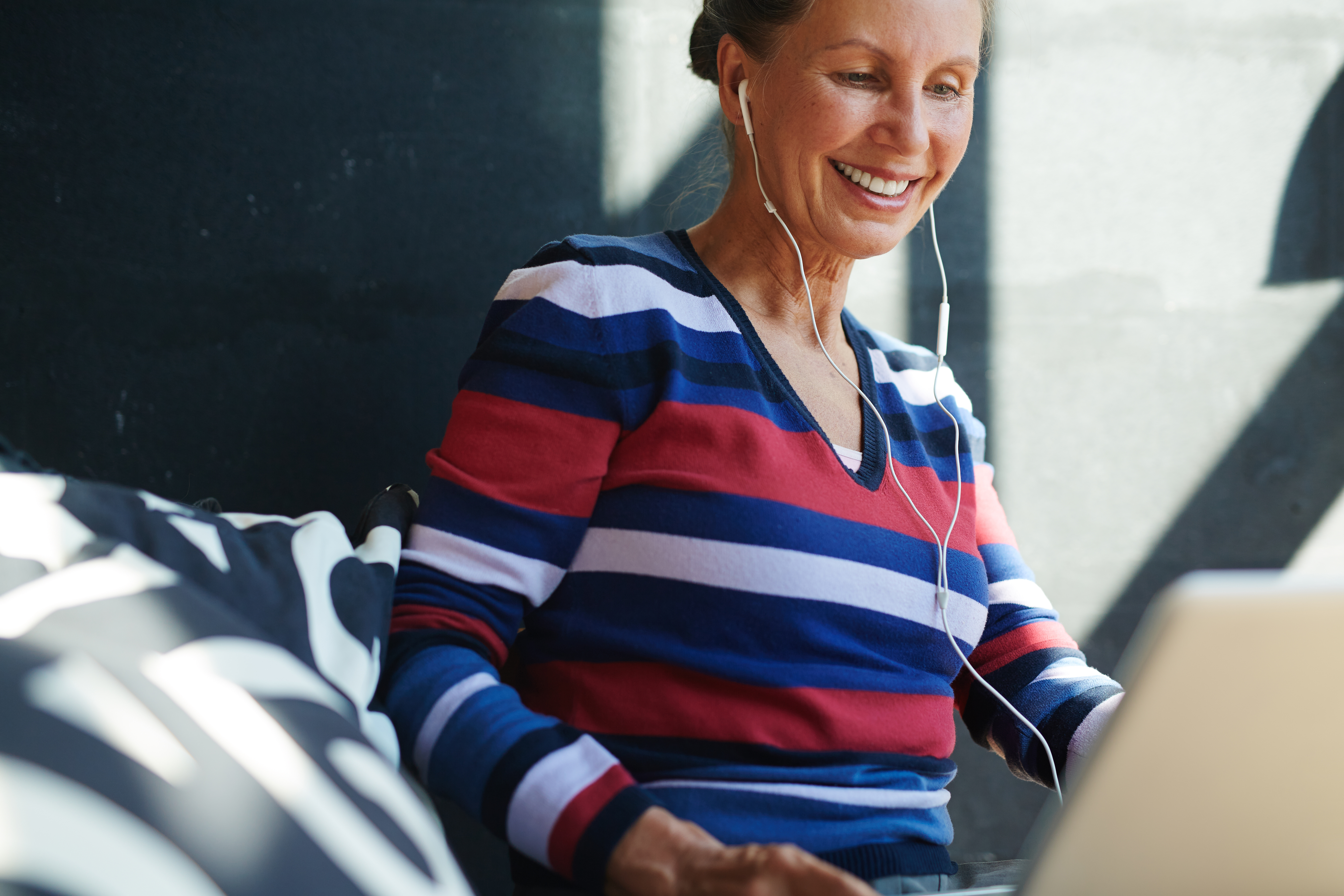 Mujer madura, sentada en el sofá con unos cascos puestos sonríe mientras mira a la pantalla de su portatil haciendo terapia online