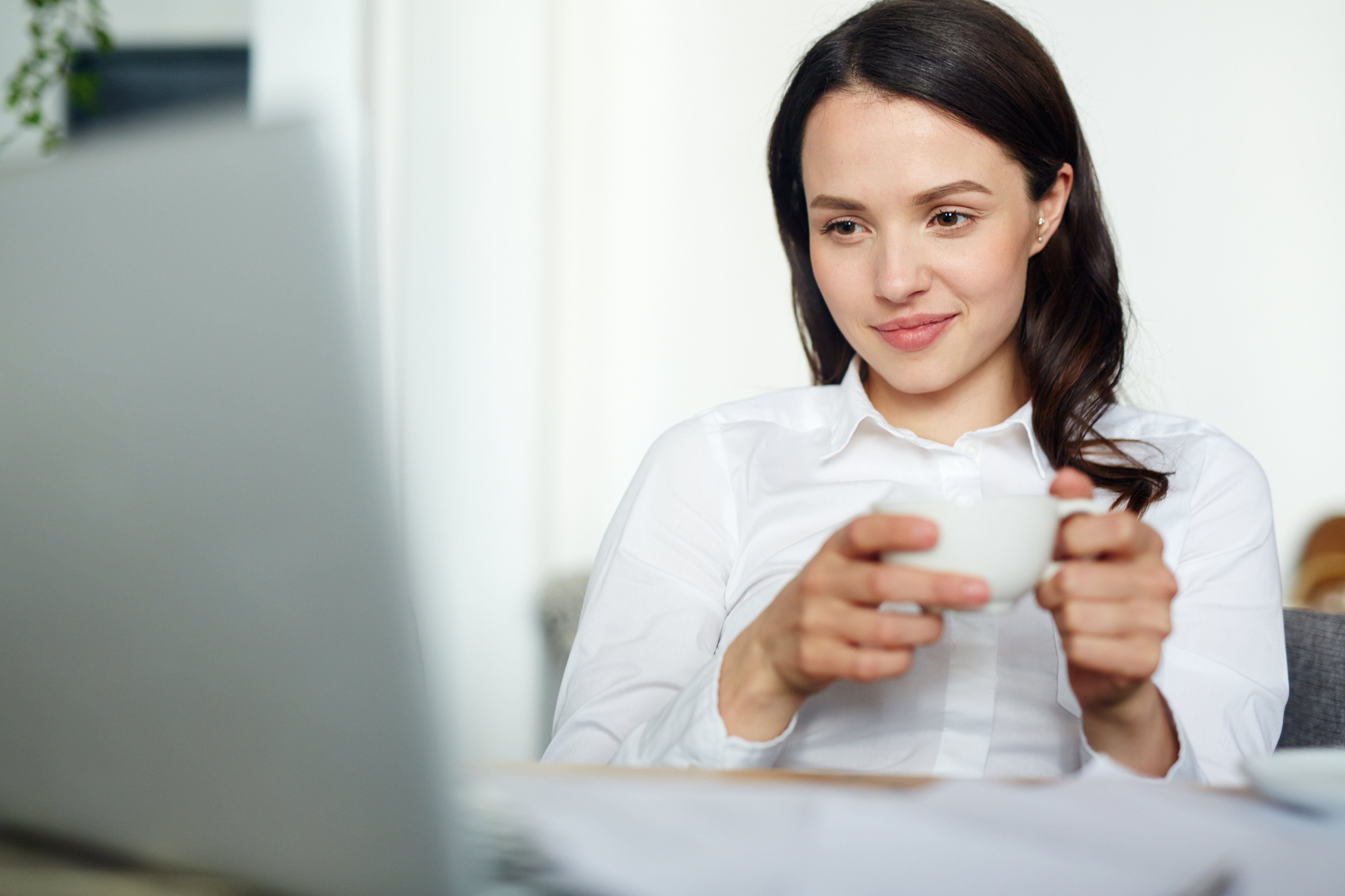 Mujer sosteniendo una taza blanca con ambas manos, sentada, mirando la pantalla de un portatil mientras esboza una sonrisa