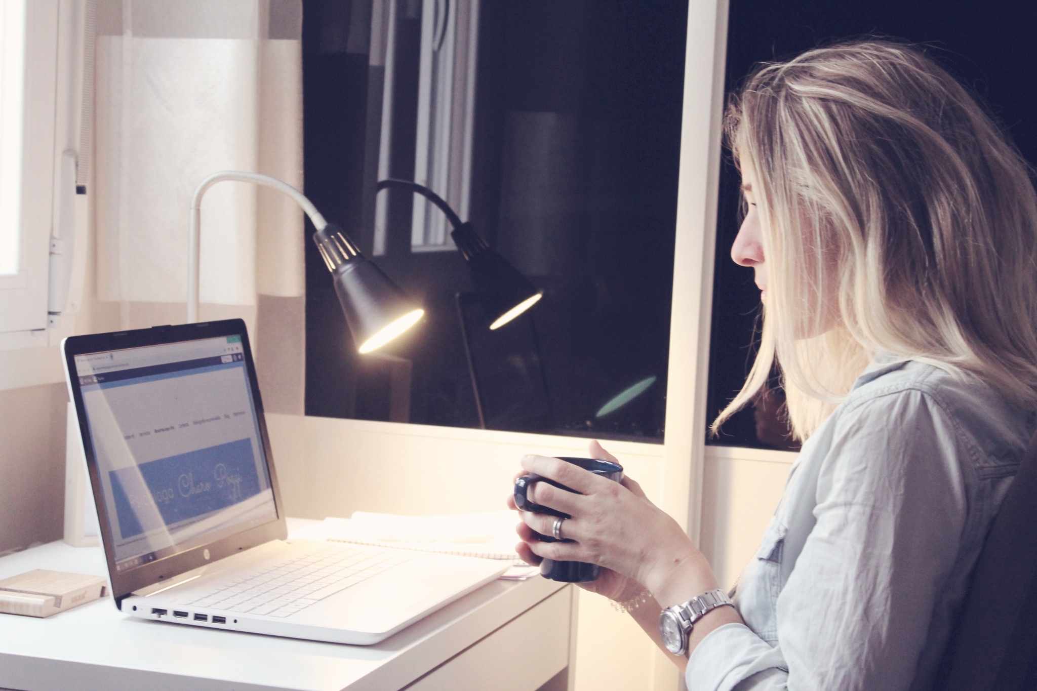 La Psicóloga Charo Poggi, mujer joven rubia, sentada mientras sostiene una taza con ambas manos, mira hacia la pantalla de su portatil donde se ve la web de psicocharopoggi.com